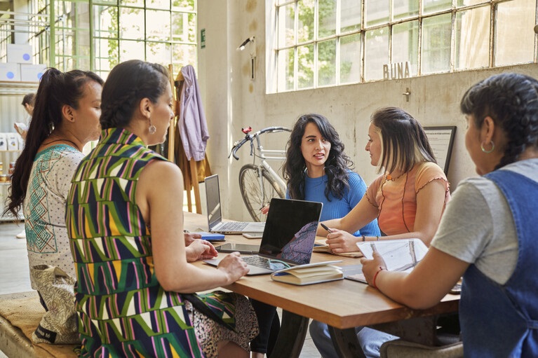 Using B2B Predictive Audiences on LinkedIn to Find Ready-to-Buy Prospects Five women around a wooden table in a bright workshop, discussing with laptops and notebooks.