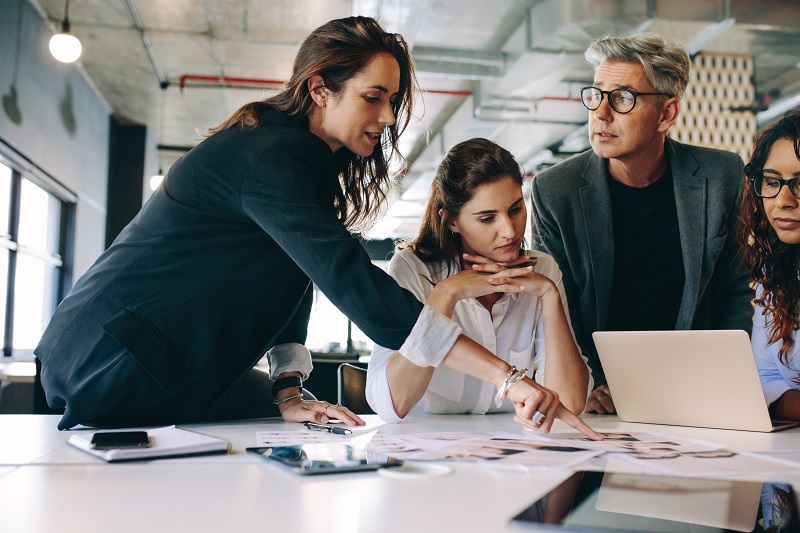 How to Date a Potential Co-Founder Before Giving Up Equity Four colleagues in a bright office lean over a table, studying photos and pointing at documents with a laptop nearby.